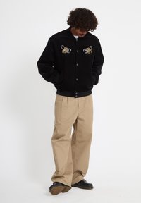 Homme regardant vers le bas, portant une veste noire avec des scorpions brodés, un pantalon large beige et des chaussures noires, debout devant un fond blanc.