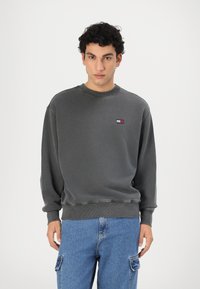 Felpa grigia con colletto e polsini a coste, con un piccolo logo ricamato. Abbinata a pantaloni cargo blu.