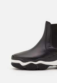 Botín Chelsea de cuero negro con panel lateral elástico y suela de goma gruesa en negro y blanco con agarre texturizado, mostrado en vista lateral en primer plano.