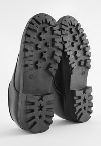 Bottes en caoutchouc noires avec des semelles texturées présentant des motifs d'étoiles, des crampons profonds et un design supérieur lisse. Taille indiquée sur la semelle.