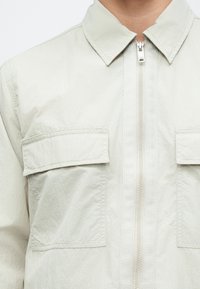 Világosszürke cipzáras dzseki hegyes gallérral, két mellkasi zsebbel és finom textúrával. Az anyag simának és könnyűnek tűnik.