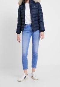 Granatowa kurtka puchowa z futrzanym kapturem, biała koszulka, jasnoniebieskie dopasowane dżinsy oraz białe trampki z czarnymi i czerwonymi akcentami.