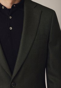 Ciemnozielona marynarka z teksturowanej tkaniny, z wyłogami szpiczastymi i pojedynczym zapięciem na guzik, noszona na czarnej koszulce polo z trzema guzikami.