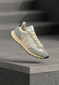 Ανοιχτό γκρι και κρεμ αθλητικό παπούτσι με μπεζ κορδόνια που αιωρείται πάνω από μια τσιμεντένια γωνία, με θολή σκάλα στο φόντο.