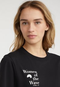 Sort sweater med "Women of the Wave" grafisk tryk i hvid, rund halsudskæring og ribbede manchetter. Blødt stof med en glat tekstur.