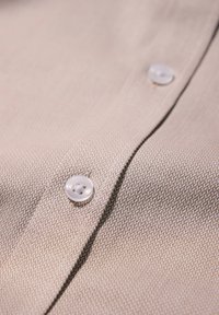 Tessuto di camicia intrecciato beige con un motivo testurizzato, caratterizzato da bottoni bianchi e un dettaglio di alette visibili.