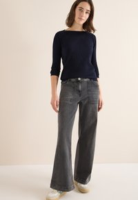 Marineblaues Rippstrickpullover mit Bootsausschnitt, kombiniert mit grauen, hoch taillierten, ausgestellten Jeans mit Fronttaschen und Knopfverschluss, getragen mit weißen Sneakers.