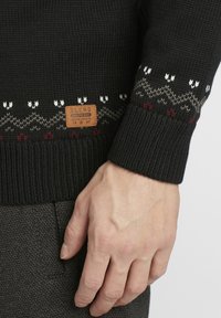 Suéter de punto negro con un diseño estampado que presenta acentos en blanco, gris y burdeos. Etiqueta cosida en la parte inferior izquierda. Puños texturizados.