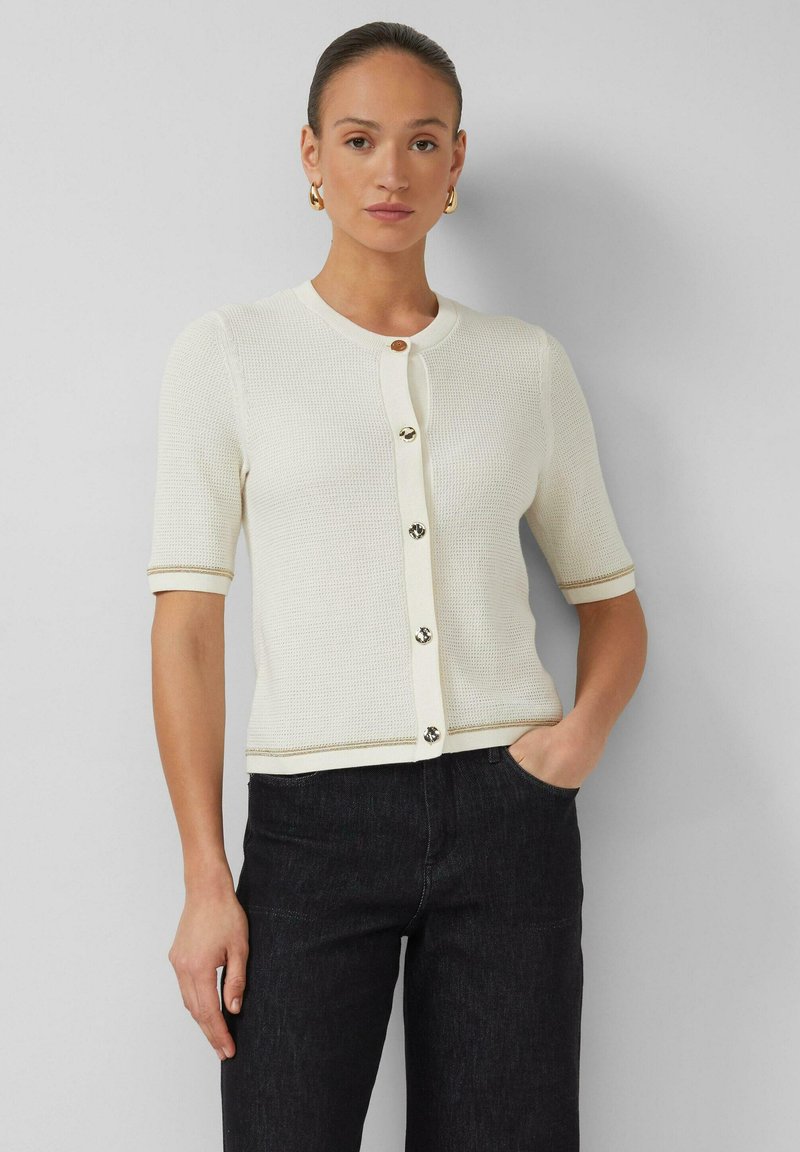 Vrouw draagt een wit kort mouwtjes gebreid vest met knopen en een zwarte broek, staand met één hand in haar zak tegen een effen achtergrond.