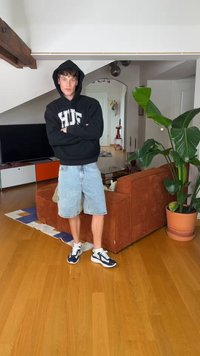 Giovane uomo con felpa nera con cappuccio e pantaloncini di jeans sta con le braccia incrociate in un soggiorno con un divano marrone e una grande pianta verde.