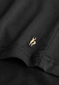 Fekete bordázott anyag közeli képe, amelyhez egy kis arany monogram részlet van csatolva a varrott varrat közelében.