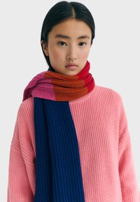 Jeune personne portant un pull en tricot rose et une écharpe côtelée multicolore en rouge, orange, rose et bleu marine, sur un fond uni.