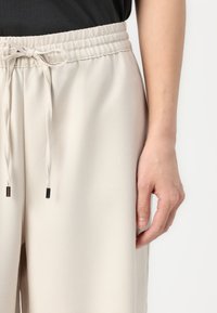Beige relaxed-fit pants with an elastic waistband, drawstring detail with metallic tips, and smooth, lightweight fabric texture.