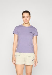 T-shirt violet à manches courtes en tissu doux, avec un logo noir Patagonia sur la poitrine gauche. Porté avec un short beige clair.