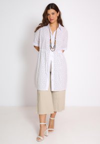 Witte short-sleeve shirt met oogjesdesign, knoopsluiting aan de voorkant en tailleband. Gecombineerd met beige wijde broek en witte block-heeled sandalen.