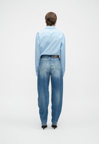 Asmuo, stovintis atsukęs nugarą, vilkintis mėlyną juostuotą marškinį, išblukusius aukšto liemens mėlynus džinsus ir juodus aukštakulnius batus ant paprasto fono.