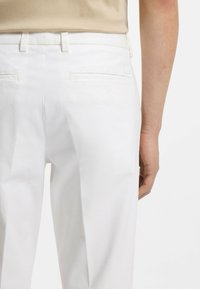 Persona vistiendo pantalones blancos a la medida y top beige, mostrando la vista de espaldas con el brazo derecho relajado a un lado.