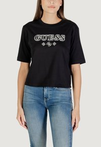 T-shirt corta nera con logo "GUESS" in argento, maniche corte, scollatura rotonda, indossata con jeans blu. Design semplice e solido con dettagli minimi.