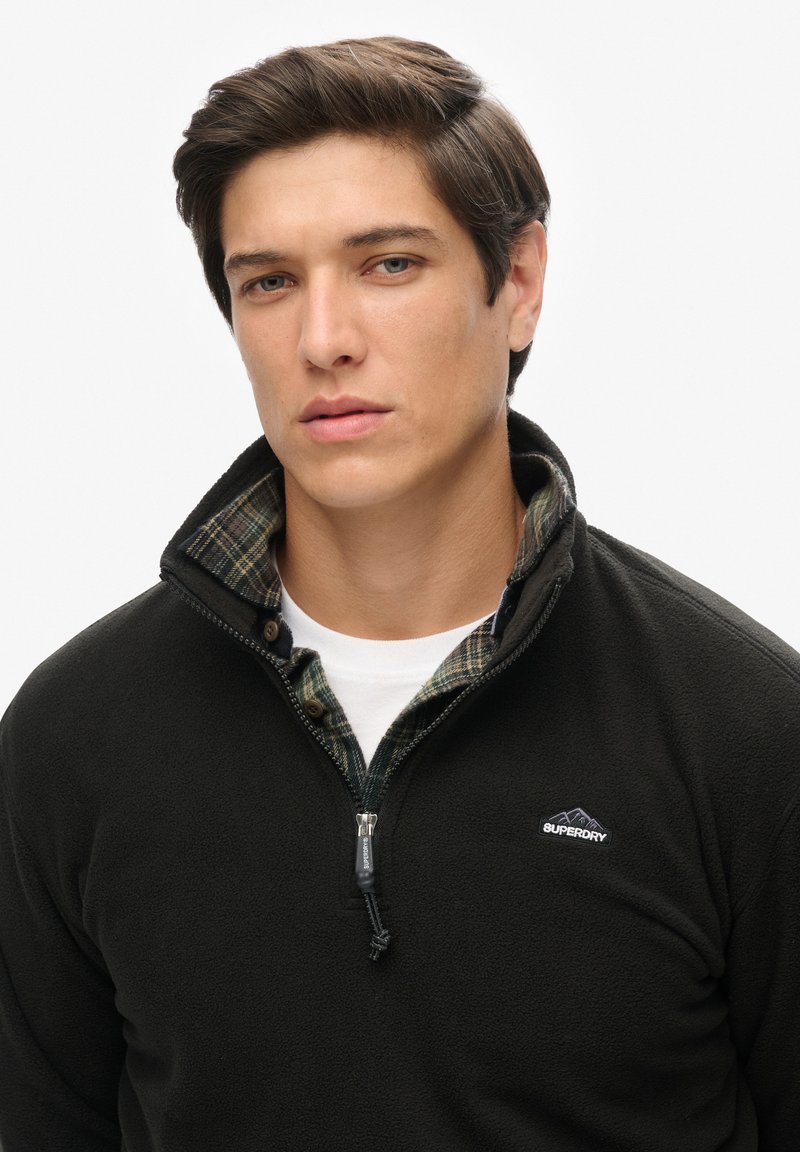 Zwarte fleece quarter-zip pullover met een kraag, voorzien van een geruite binnenvoering en een klein logo op de borst. Soepele textuur en zacht materiaal.