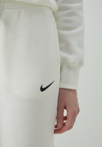 Bijele pamučne jogger hlače s mekim tekstilom, s crnim Nike swoosh logom na bedru. Ruka se oslanja na tkaninu, ističući dizajn.