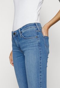 Närbild av en person som bär tajta blå jeans och en vit topp, med en hand i framlommen, mot en neutral bakgrund.
