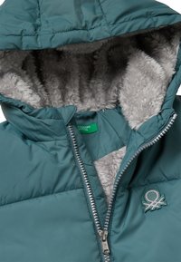 Teal isolierte Jacke mit einem Kunstpelz-Futter, ausgestattet mit einem Reißverschluss und einem gestickten Logo auf der Brust. Satin-Finish, weiche Textur.