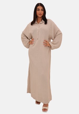 Une femme pose avec confiance dans une robe longue beige clair à manches longues avec un col, assortie à des chaussures ouvertes élégantes.