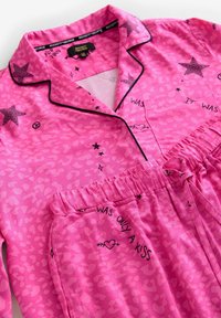 Set de pijamale roz cu imprimeu leopard, cu accente de stele negre și text. Bluza are guler și închidere cu nasturi; pantalonii au talie elastică.