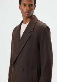 Bruine dubbelgeknoopte jas met visgraatstructuur, reverskraag met inkeping en twee voorzakken. Gemaakt van duurzaam stof; voorzien van donkere knopen.