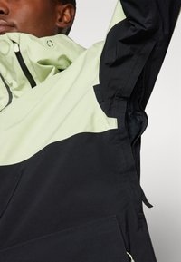 Giacca nera e verde chiaro con una finitura testurizzata. Presenta un collo alto, tasche laterali e un design con pannelli a contrasto. Inclusi zip funzionali.