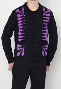 Svart stickad cardigan med stora lila blommönster framme och på fickorna. Den har en knappstängning och krage.