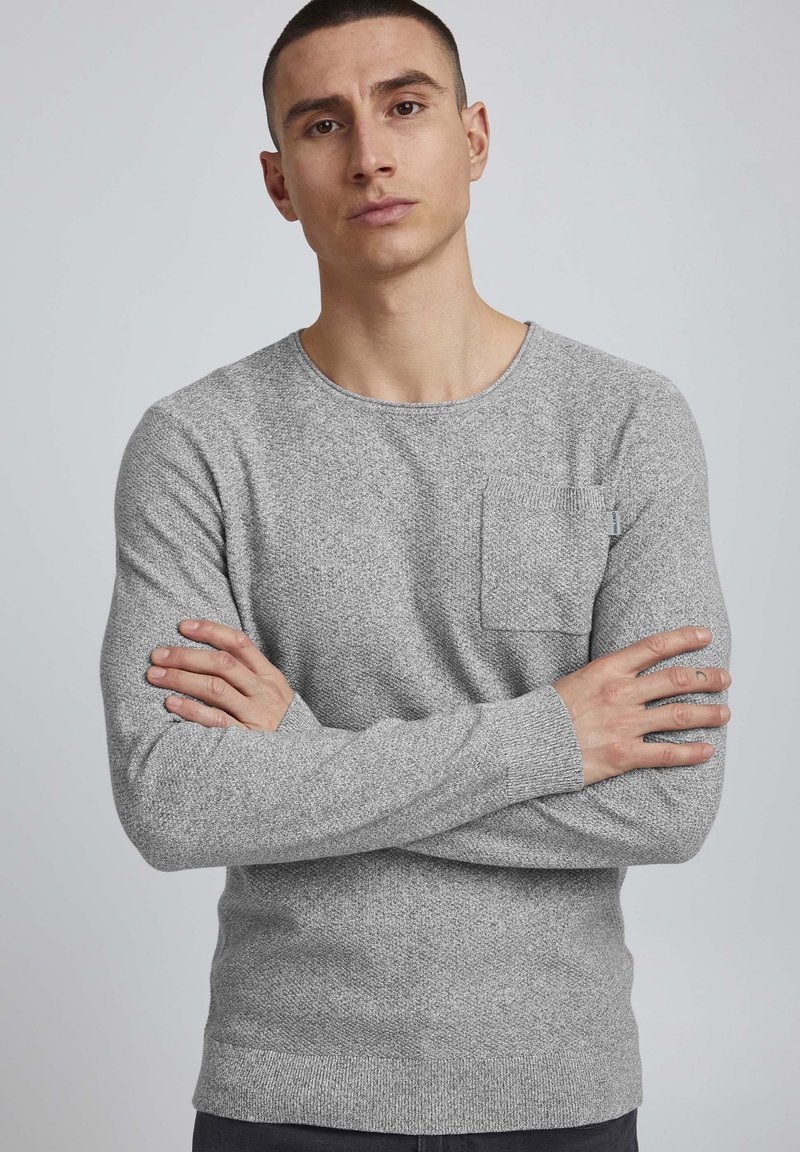 Pull en tricot gris avec un col rond, des manches longues et une petite poche poitrine ; motif texturé et poignets et ourlet côtelés.