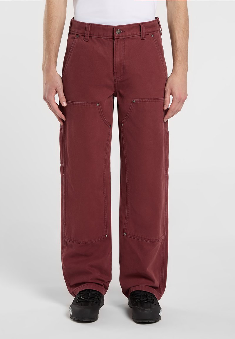 Uomo con pantaloni cargo rossi scuri e scarpe nere, che sta in piedi contro uno sfondo bianco semplice, con le mani appoggiate accanto ai pantaloni.