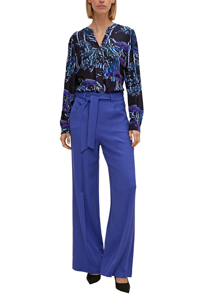Femme portant un chemisier manches longues noir et bleu à motifs, rentré dans un pantalon bleu taille haute, jambes larges, avec une ceinture en tissu.
