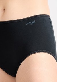 Primo piano di una persona che indossa mutande nere a vita alta con il logo "sloggi" sulla cintura, su uno sfondo neutro.