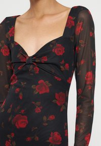 Robe noire avec motif de roses rouges, manches longues transparentes et encolure carrée avec détail froncé au niveau de la poitrine.