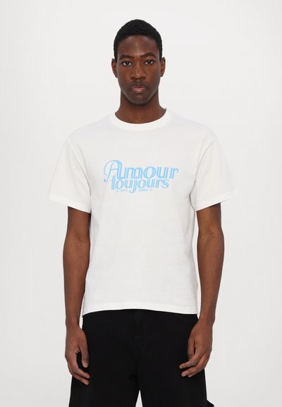 White cotton t-shirt featuring blue text "Amour Toujours" with a curved font. Short sleeves and crew neck design. Worn with black pants.