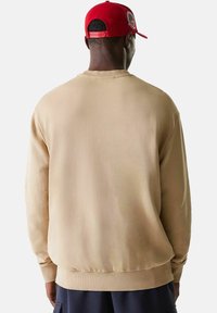 Mies, který má na sobě béžovou mikinu a červenou čepici, obrácený zády k jednoduchému bílému pozadí.