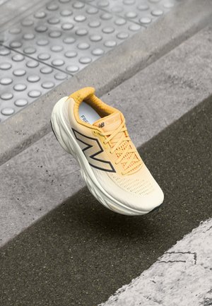 Chaussure de course New Balance jaune et blanche flottant au-dessus d'un trottoir gris texturé et d'un bord avec revêtement tactile à proximité.