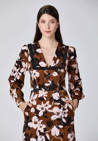 Robe florale en tissu marron ornée de grandes fleurs blanches et noires. Elle possède des manches longues, un col volanté et des poches latérales.