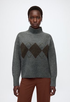 Femme portant un pull à col roulé gris avec un motif losange foncé et un pantalon marron sur fond blanc uni.