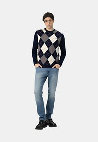 Marineblaues Argyle-Pullover mit weißen, grauen und roten Rauten; gerippte Bündchen; kombiniert mit hellblauen Jeans und schwarzen Schuhen.