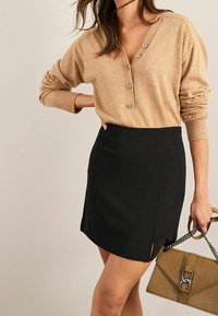 Femme portant un pull boutonné beige et une jupe noire fendue sur le côté, tenant un sac à main texturé couleur tabac avec une bandoulière en chaîne.