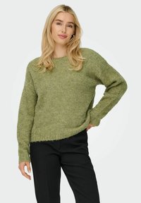 Groene gebreide trui met een wijde halslijn en lange mouwen, met een zachte textuur en een iets verkorte zoom, gecombineerd met zwarte broeken.