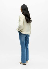 Cremefarbener Strickpullover mit texturierten Verzierungen auf der linken Schulter, kombiniert mit hellblauen weit geschnittenen Jeans und beigen Slippern.