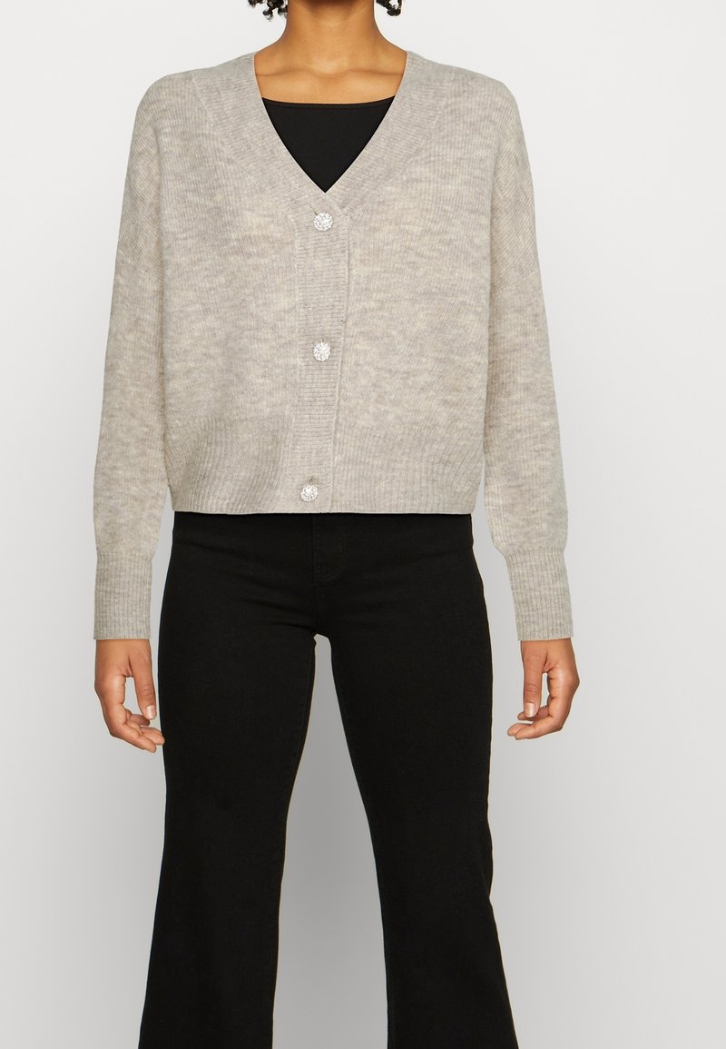 Personne portant un cardigan tricoté gris clair avec trois boutons scintillants sur un haut noir et un pantalon noir sur un fond uni.