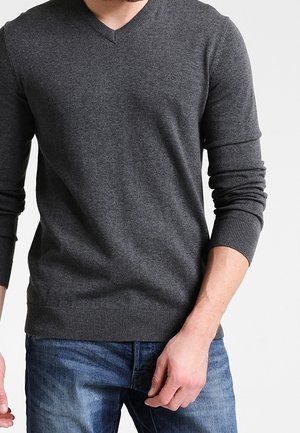 Homme portant un pull gris foncé en V et un jean bleu, debout avec une main près de sa poche devant un fond uni.