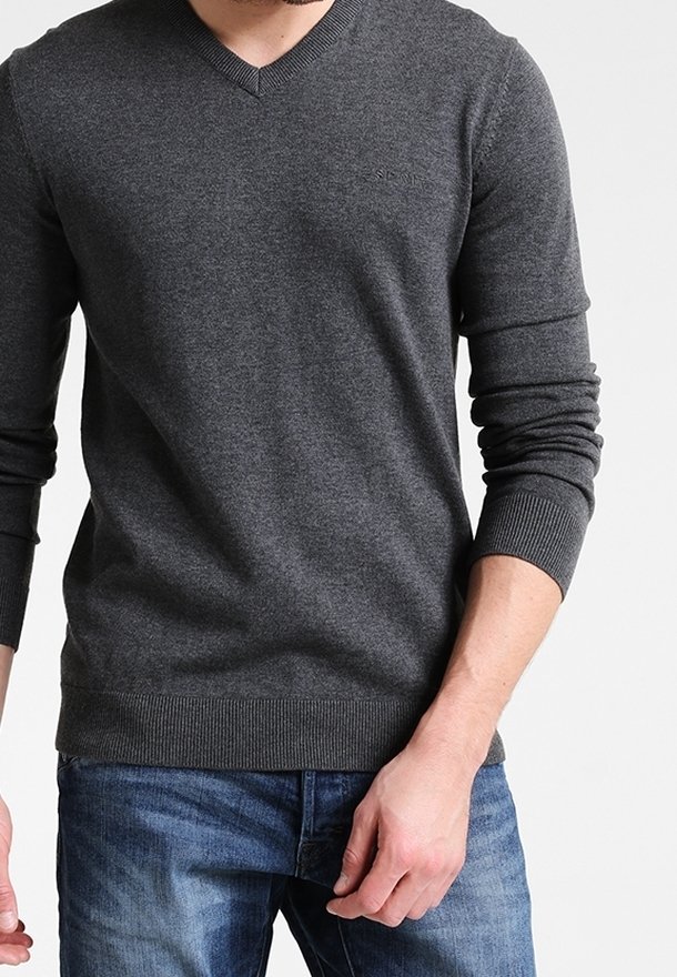 Homme portant un pull gris foncé en V et un jean bleu, debout avec une main près de sa poche devant un fond uni.