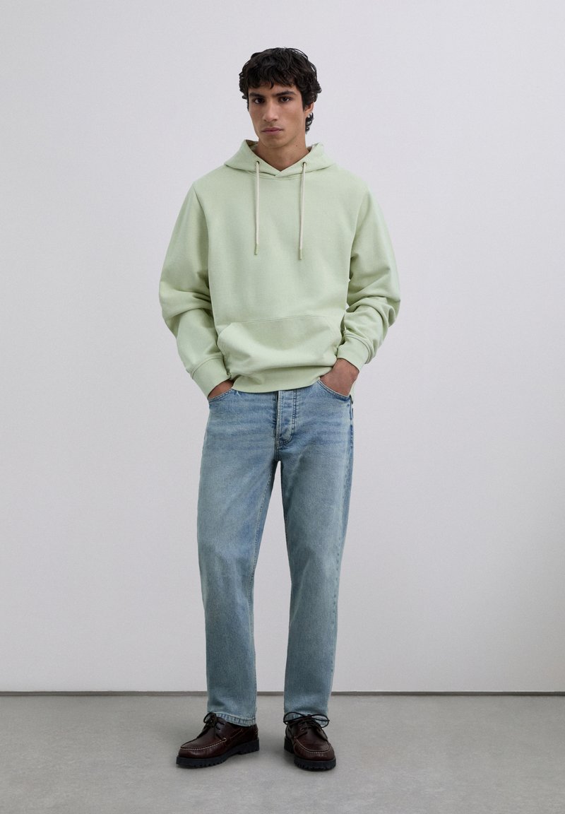 Man standing with hands in pockets wearing a light green hoodie, blue jeans, and brown leather shoes against a plain backdrop.