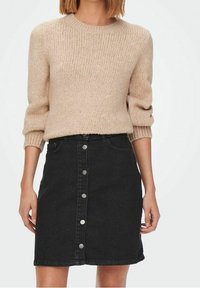 Femme portant un pull en maille beige aux manches retroussées et une jupe en jean noire taille haute avec des boutons sur le devant.